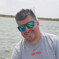 Dave Alvarado, smiling, in sunglasses on a lake, with a sailboat behind him.
